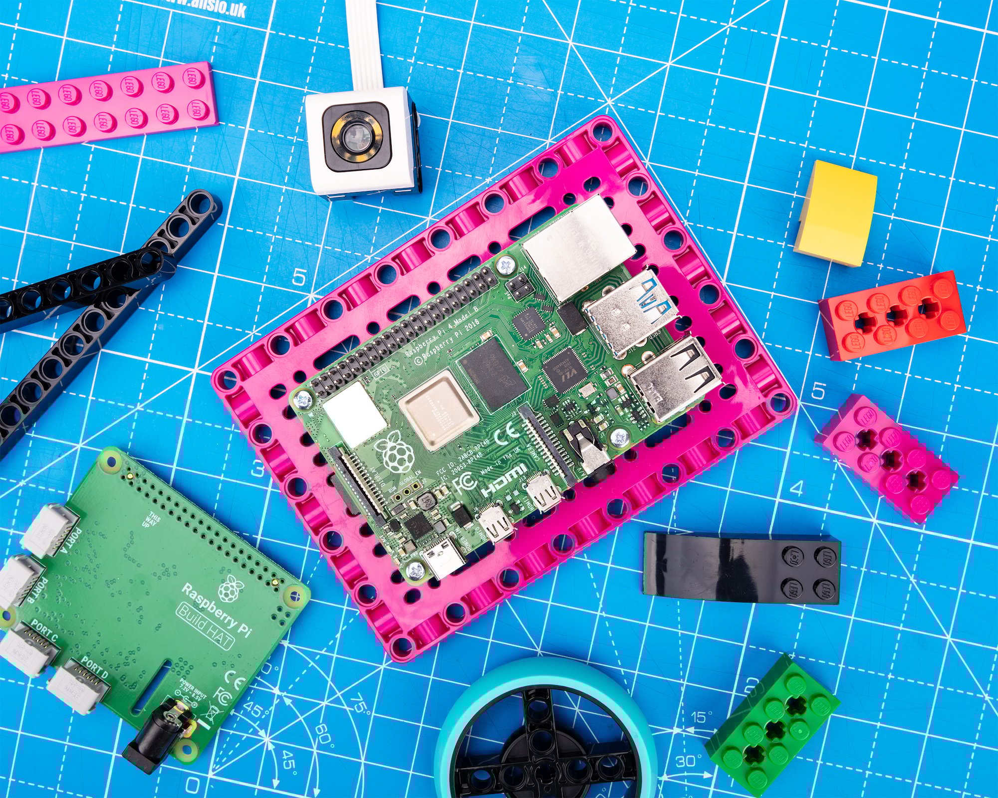 Top-down photo of a Raspberry Pi mounted on a large, flat, bright pink LEGO element which we now know is the LEGO Maker Plate. A varied selection of other LEGO elements, along with a Build HAT, are scattered around it.